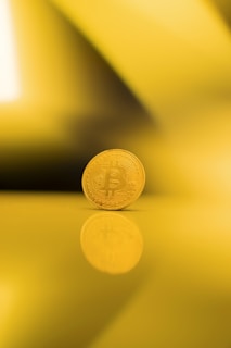 A single gold-colored Bitcoin standing upright on a reflective surface, casting a clear reflection. The background consists of various shades of yellow, creating an abstract and warm ambiance.