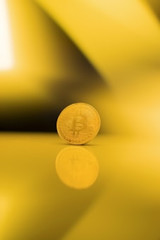 A single gold-colored Bitcoin standing upright on a reflective surface, casting a clear reflection. The background consists of various shades of yellow, creating an abstract and warm ambiance.