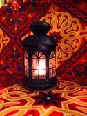 An ornate black lantern with glass panels is glowing with light from within. It rests on an intricately patterned red and orange fabric backdrop that features symmetrical, floral, and geometric designs.