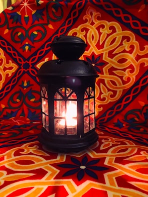 An ornate black lantern with glass panels is glowing with light from within. It rests on an intricately patterned red and orange fabric backdrop that features symmetrical, floral, and geometric designs.