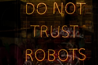 A neon sign displaying the words 'DO NOT TRUST ROBOTS' with a dark background and various reflections. The letters are illuminated in a warm yellowish-orange color, creating a stark contrast against the dim setting.