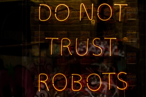 A neon sign displaying the words 'DO NOT TRUST ROBOTS' with a dark background and various reflections. The letters are illuminated in a warm yellowish-orange color, creating a stark contrast against the dim setting.