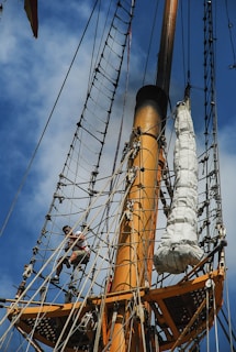 A person is climbing a series of ropes on the mast of a tall ship. The mast is painted orange, and the ropes form a network around it. A large bundle is secured with ropes to the right of the climber. The sky is partially visible and features scattered clouds.