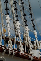 A close-up view of a ship's rigging, featuring a series of ropes, pulleys, and white hardware against a background of cloudy sky. The ropes are tightly secured, with white and wooden elements prominently displayed, suggesting part of a tall ship's structure.