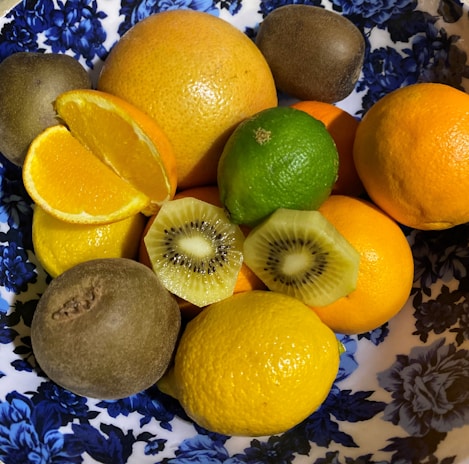 A variety of fruits, including kiwi slices, lemons, oranges, limes, and whole kiwis, are arranged in a decorative bowl with a blue floral pattern. The fruits display a range of vibrant colors and textures.