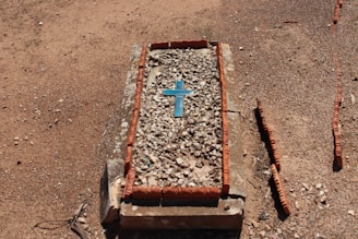 A grave covered with small stones features a blue cross in the center. The surrounding ground is gravelly and dry, with a border of reddish bricks outlining the grave.