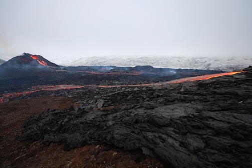 A volcanic landscape featuring an active volcano emitting lava and smoke. The foreground is filled with dark volcanic rock, creating a stark contrast against the vibrant orange and red hues of the flowing lava. A misty atmosphere envelops the scene, with distant mountains capped by snow or ash visible in the background.