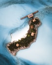A small, lush tropical island is surrounded by vibrant turquoise waters. The island features several small, thatched-roof structures connected by a wooden boardwalk. The landscape is dotted with dense greenery, and a sandy beach area is visible. A solar panel is mounted on one of the structures, and the water around the island is clear, revealing some underwater details.