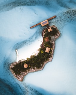 A small, lush tropical island is surrounded by vibrant turquoise waters. The island features several small, thatched-roof structures connected by a wooden boardwalk. The landscape is dotted with dense greenery, and a sandy beach area is visible. A solar panel is mounted on one of the structures, and the water around the island is clear, revealing some underwater details.