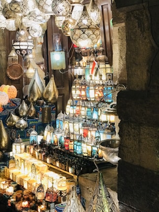 A market stall is filled with an array of colorful, intricately designed lanterns and lamps. The lanterns display a variety of patterns and colors, including blue, red, and gold. Metalware is also visible, reflecting the illumination from the lamps. The setting exudes an atmosphere of cultural richness, with traditional Middle Eastern designs prominently featured.