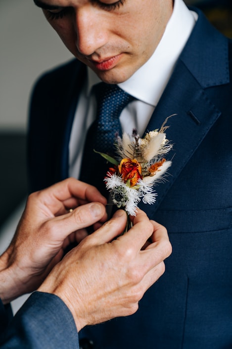 A man is wearing a navy blue suit and a white shirt, with a detailed floral boutonniere being adjusted on his lapel. The boutonniere includes dried flowers and greenery, and there is a textured blue tie visible.