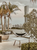 A rooftop terrace featuring modern outdoor furniture including a hammock, chairs, and tables. Palm trees and large buildings are visible in the background under a cloudy sky. Lush greenery frames the area, adding a tropical ambiance.