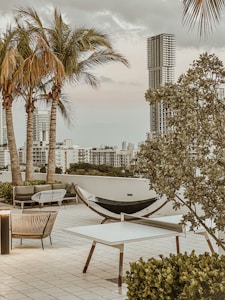 A rooftop terrace featuring modern outdoor furniture including a hammock, chairs, and tables. Palm trees and large buildings are visible in the background under a cloudy sky. Lush greenery frames the area, adding a tropical ambiance.