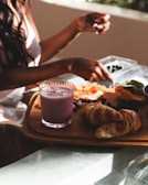 A breakfast setting on a wooden tray is adorned with a glass of purple smoothie, croissants, salmon slices, avocado, and blueberries. Sunlight casts a warm glow over the food as a person reaches for a piece of fruit.