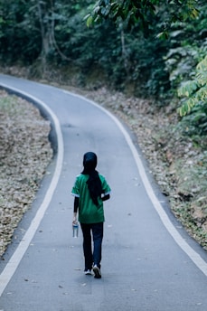 A person wearing a black headscarf and green shirt walks alone along a winding road surrounded by dense green foliage. The road is lined with fallen leaves, creating an atmosphere of solitude and nature.