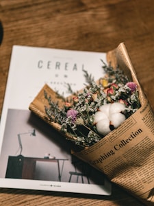 A bouquet of dried flowers wrapped in newspaper lies on top of an open magazine. The magazine features a minimalist design with a desk lamp visible. The wooden surface adds a warm, rustic feel to the composition.