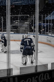 A hockey player wearing full gear stands on an ice rink, facing away. The player is in a dark blue uniform with the name Yeryomenko on the back. Another player and an empty stadium with advertisements on the surrounding boards are visible in the background.