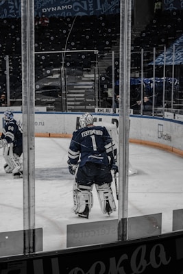 A hockey player wearing full gear stands on an ice rink, facing away. The player is in a dark blue uniform with the name Yeryomenko on the back. Another player and an empty stadium with advertisements on the surrounding boards are visible in the background.
