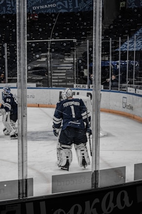 A hockey player wearing full gear stands on an ice rink, facing away. The player is in a dark blue uniform with the name Yeryomenko on the back. Another player and an empty stadium with advertisements on the surrounding boards are visible in the background.