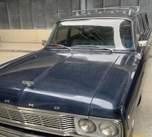 A vintage Ford car is parked indoors, featuring a glossy dark blue exterior with a distinct front grille and classic design elements. The car appears to be well-maintained, showcasing the elegant curves typical of older automotive models. A roof rack is visible on top, and the setting includes a concrete floor and metal wall panels in the background.