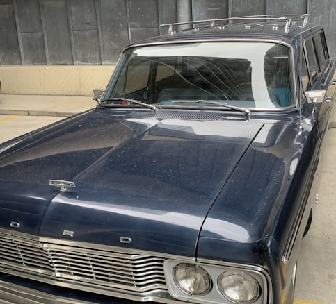 A vintage Ford car is parked indoors, featuring a glossy dark blue exterior with a distinct front grille and classic design elements. The car appears to be well-maintained, showcasing the elegant curves typical of older automotive models. A roof rack is visible on top, and the setting includes a concrete floor and metal wall panels in the background.