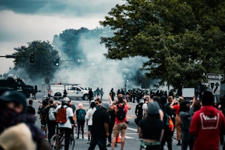 A group of people gathered on a street, possibly participating in a protest. There is a large presence of police officers in riot gear facing the crowd, with smoke or tear gas in the air. Vehicles, including police vans and other cars, are scattered throughout the scene. Some individuals are taking photos or holding signs.