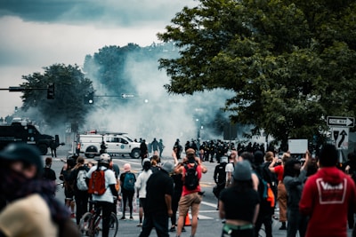 A group of people gathered on a street, possibly participating in a protest. There is a large presence of police officers in riot gear facing the crowd, with smoke or tear gas in the air. Vehicles, including police vans and other cars, are scattered throughout the scene. Some individuals are taking photos or holding signs.