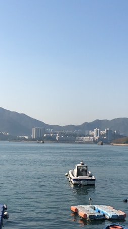 A small motorboat is positioned on a calm body of water with a mountainous backdrop. In the distance, a cluster of modern buildings lines the shoreline. The sky is clear and blue, suggesting a bright day.