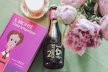 A purple book with a colorful illustration on the cover titled 'Cime tempestose' by E. Brontë is placed next to a dark bottle labeled '2 Sorelle' on a textured green surface. Nearby, a delicate flower arrangement with soft pink peonies and a lit white candle on a decorative saucer add to the aesthetic.