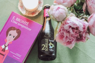 A purple book with a colorful illustration on the cover titled 'Cime tempestose' by E. Brontë is placed next to a dark bottle labeled '2 Sorelle' on a textured green surface. Nearby, a delicate flower arrangement with soft pink peonies and a lit white candle on a decorative saucer add to the aesthetic.