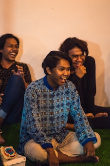 Three people are sitting on the floor, laughing and enjoying each other's company. The person in the center is wearing a blue patterned shirt and beige pants, while the other two are dressed in dark clothing. A piece of cake is visible on the grass next to them.