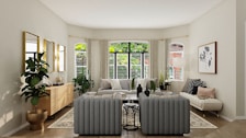 A well-furnished living room with a modern, minimalist aesthetic. The space features a large window framed by light curtains, allowing natural light to flood the room. There is a gray tufted sofa set, a wooden sideboard, several potted plants, and a large framed artwork on the wall. The color palette is neutral with green and wooden accents.