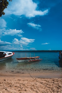 A tranquil beach scene with a small boat anchored close to the shore, clear turquoise water, and a bright blue sky with scattered white clouds. The sandy beach is visible in the foreground, and a larger boat is partially in view on the left side.