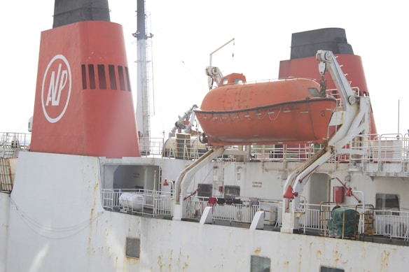 A large vessel with two prominent smokestacks featuring a red design and logo. An orange lifeboat is suspended on the deck with metal arm supports. The ship's exterior is white and slightly weathered, with railings and other equipment visible.