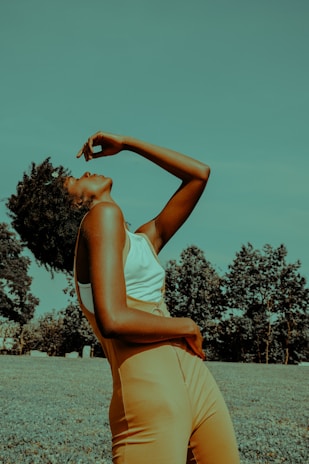 A person stands in a relaxed pose on a grassy area surrounded by trees. Their head is tilted back and one arm is raised above their head, while the other arm rests on their midsection. The person is wearing a white top and high-waisted mustard-colored pants. The image has a warm, slightly surreal color tone.