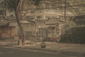 A quiet street scene featuring a small makeshift stall selling goods like snacks and beverages. The stall is situated on a sidewalk with a tree providing shade. Nearby, there are red plastic stools and various items hanging from the stall. A large stone ball is placed on the pavement close to the road. Surrounding structures appear to be old and slightly dilapidated, adding a rustic character to the setting.
