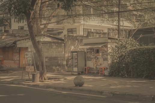A quiet street scene featuring a small makeshift stall selling goods like snacks and beverages. The stall is situated on a sidewalk with a tree providing shade. Nearby, there are red plastic stools and various items hanging from the stall. A large stone ball is placed on the pavement close to the road. Surrounding structures appear to be old and slightly dilapidated, adding a rustic character to the setting.