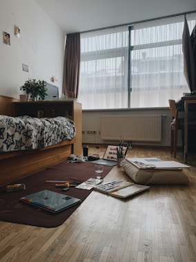 A cozy bedroom with a large window covered by sheer curtains, allowing diffused light to enter. The room features a bed with a patterned comforter, a wooden desk with a plant and some personal items. On the floor, there is an open laptop, art supplies like paints and brushes, along with sketchbooks and papers. A cushion is also present on the wooden floor, suggesting a casual, creative atmosphere.