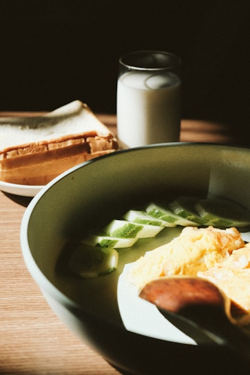 A close-up view of a breakfast setting featuring a rustic bowl with slices of cucumber and a portion of scrambled eggs, accompanied by a wooden spoon. In the background, there's a stack of sliced bread and a glass of milk, all illuminated by soft natural light.