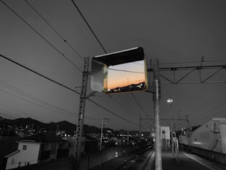 A grayscale image of a railway station features a vibrant sunset reflection in a mirror suspended above. The station is empty, with visible train tracks, overhead power lines, and small structures and houses in the background. The contrast between the monochrome surroundings and the colorful sunset reflection creates a striking visual effect.