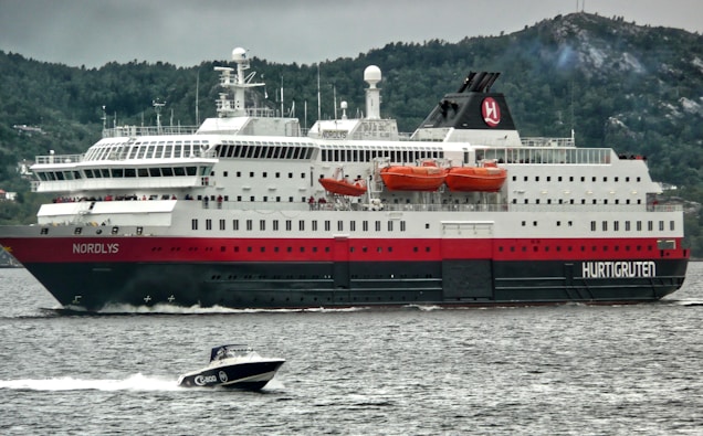 A large passenger ferry is navigating through a body of water, accompanied by a smaller motorboat in the foreground. The ferry is predominantly white with red and black accents, and has several orange lifeboats mounted on its side. It is cruising near a forested landscape with hills in the background under a cloudy sky.