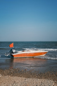 A small motorboat with an orange hull and an outboard motor is positioned at the edge of a beach, where the waves meet the shore. The boat features a red flag at its rear. Surrounding it, the sea extends to the horizon under a clear blue sky, with a sandy, pebble-strewn shoreline in the foreground.