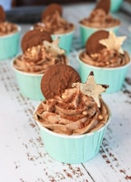 A collection of cupcakes with light blue wrappers is arranged on a textured white surface. Each cupcake is topped with swirls of chocolate frosting, a round biscuit, edible gold stars, and small decorative gold balls.