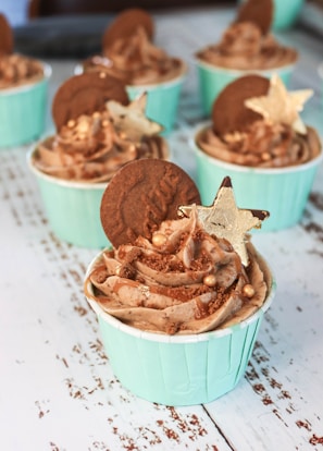 A collection of cupcakes with light blue wrappers is arranged on a textured white surface. Each cupcake is topped with swirls of chocolate frosting, a round biscuit, edible gold stars, and small decorative gold balls.