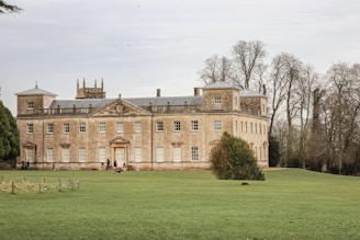 A large, historic manor house with a stately appearance is set amidst a vast, well-maintained lawn. The building features symmetrical design elements with numerous windows and decorative architectural details. Winters trees are visible in the background, adding to the serene atmosphere of the grounds.