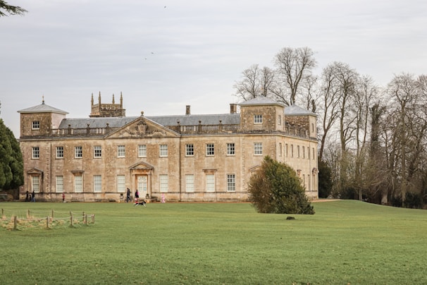 A large, historic manor house with a stately appearance is set amidst a vast, well-maintained lawn. The building features symmetrical design elements with numerous windows and decorative architectural details. Winters trees are visible in the background, adding to the serene atmosphere of the grounds.