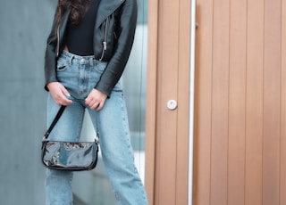 woman in black leather jacket and blue denim jeans standing beside brown wooden door