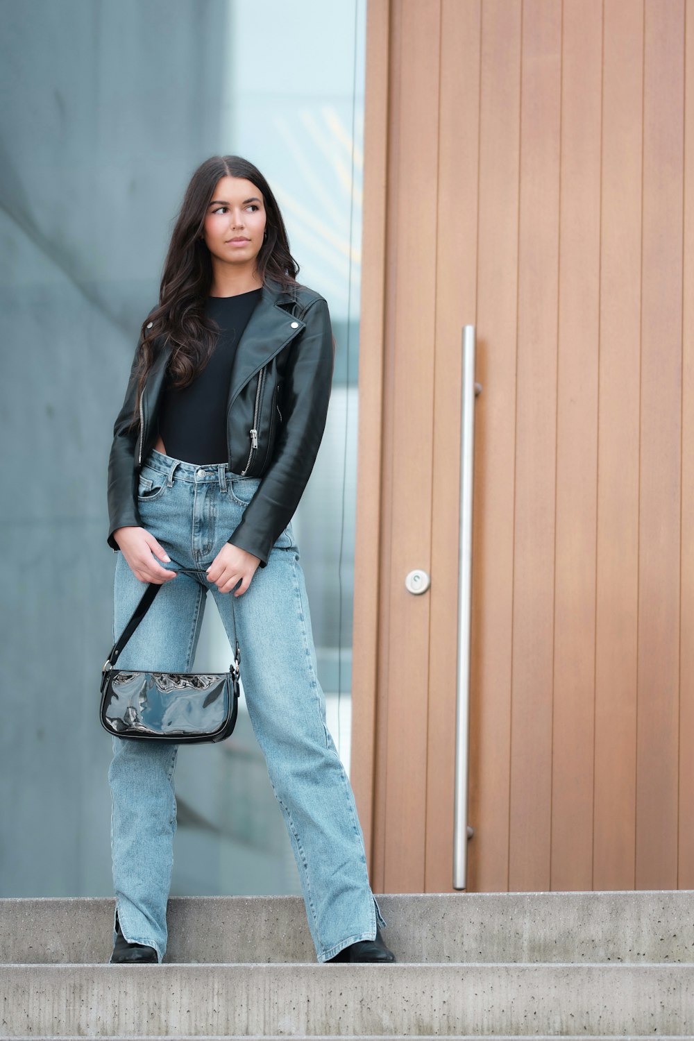 woman in black leather jacket and blue denim jeans standing beside brown wooden door