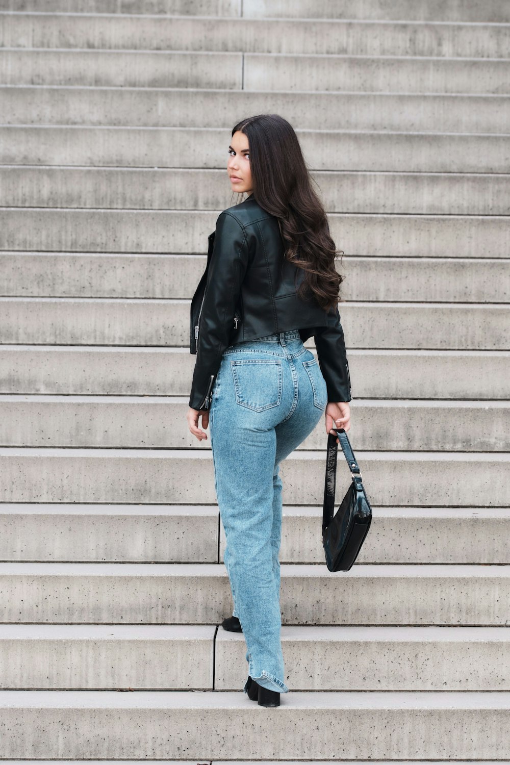 woman in black jacket and blue denim jeans standing beside white wall