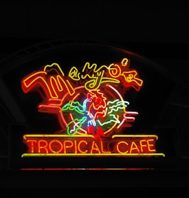 A vibrant neon sign displays a tropical theme with bright, colorful lights. The sign includes the words 'Tropical Cafe' prominently, accented by a stylized parrot and palm leaves in neon colors. The overall design suggests a lively and inviting atmosphere associated with tropical or island vibes.